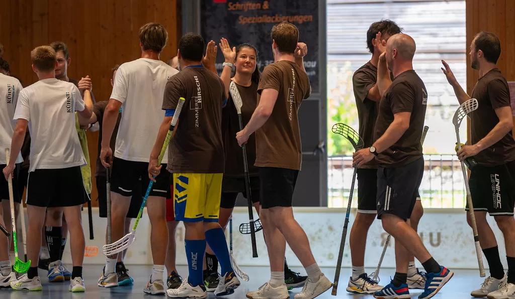 Zwei Unihockey-Teams klatschen sich nach dem Spiel ab – zu sehen sind Spielende in weißen und braunen Shirts mit Unihockeyschlägern. In der Mitte hebt eine Spielerin der Zimmerei Hirschi AG die Hand zum High-Five, rundherum ist sportliche Kameradschaft zu spüren.