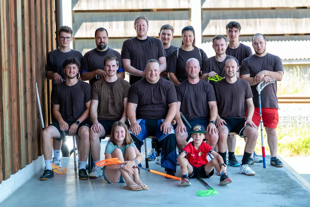 Eine fröhliche Gruppe von 17 Personen – Männer, eine Frau und zwei Kinder – posiert jubelnd für ein Gruppenfoto. Alle tragen Sportkleidung, viele einheitlich braune T-Shirts, und einige halten Unihockeyschläger in der Hand. Die Stimmung ist ausgelassen, viele recken die Arme in die Höhe. Im Vordergrund sitzen zwei Kinder mit hochgestreckten Armen, im Hintergrund ist ein Holzgebäude und Sonnenlicht zu sehen – ein sportlicher Team-Moment voller Energie.