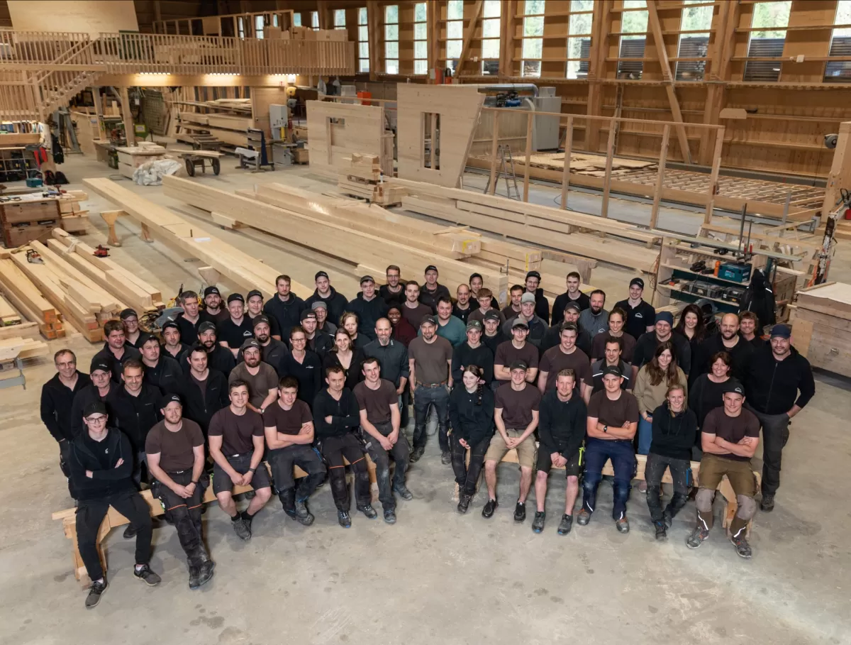 Alle Menschen, die für die Truberholz AG arbeiten auf einem Bild. Das Bild wurde in der Truberholzhalle erstellt. Die Personen stehen zwischen Balken verteilt. Das Holz und die Halle ist ersichtlich. Hinten sieht man die Truberholzanlage. Der Frauenanteil ist auch sehr gut zu sehen.