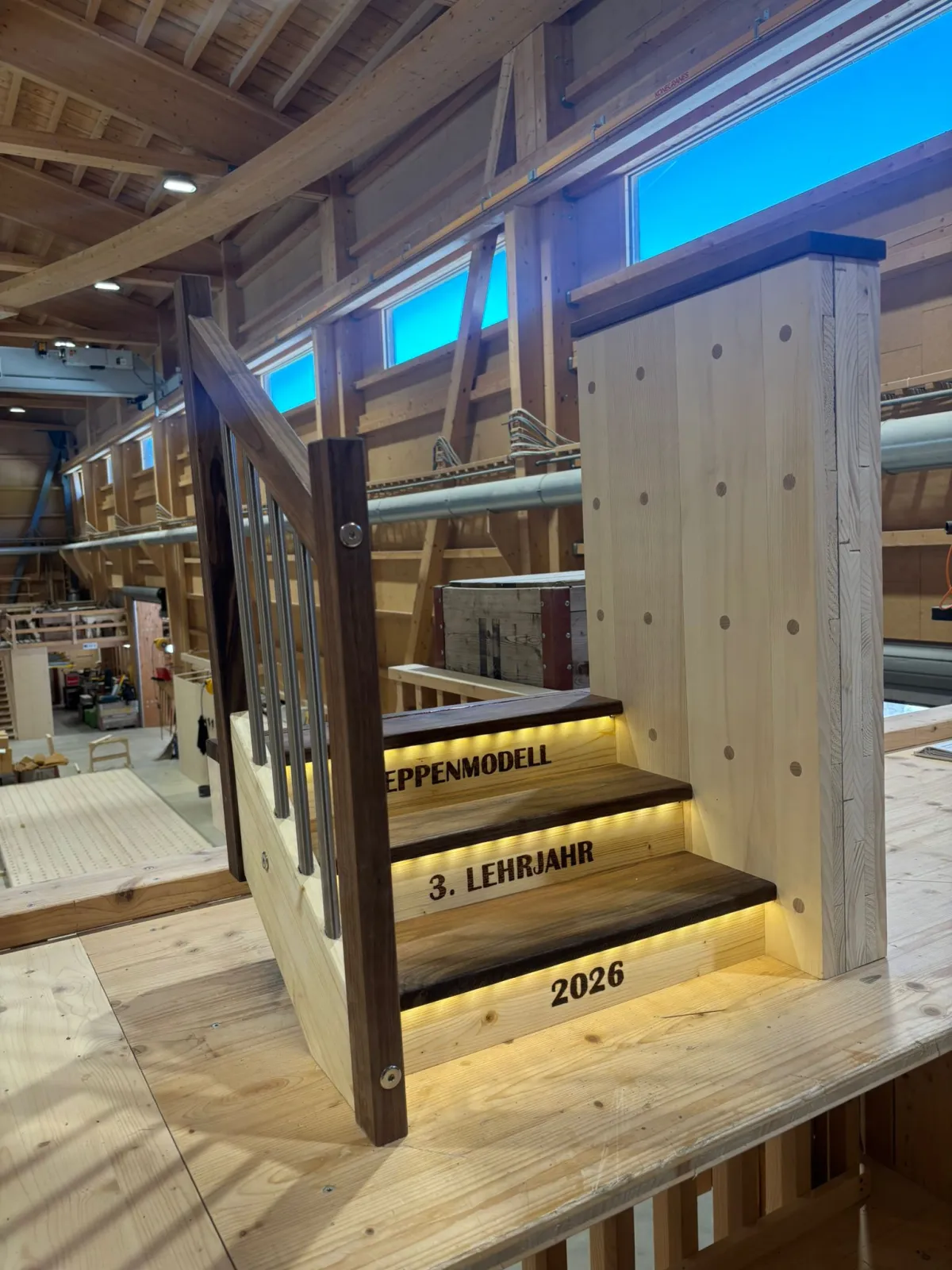 Eine Lernende im Holzbau lehnt lächelnd an einem Holzgeländer und steht neben einem beleuchteten Treppenmodell mit der Aufschrift ‚Treppenmodell, 3. Lehrjahr, 2026‘. Die Szene befindet sich in einer grossen, modernen Zimmerei-Werkhalle mit sichtbarer Holzkonstruktion, Dachbalken und verschiedenen Holzbauelementen im Hintergrund.