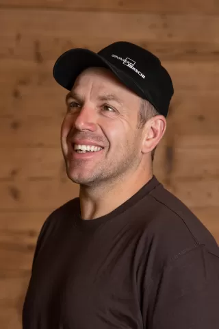 Mitarbeiterportrait / Foto mit braunem T-Shirt oder schwarzem Polo Shirt mit Zimmerei Hirschi Logo. Strahlend und lachend nach oben schauend oder direkt in die Kamera.