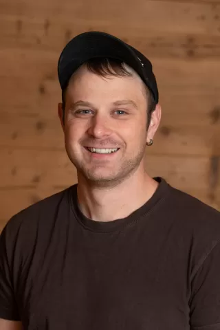Mitarbeiterportrait / Foto mit braunem T-Shirt oder schwarzem Polo Shirt mit Zimmerei Hirschi Logo. Strahlend und lachend nach oben schauend oder direkt in die Kamera.
