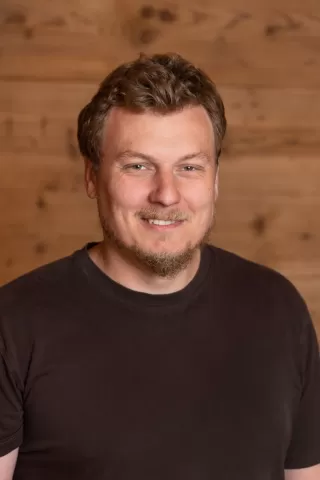 Mitarbeiterportrait / Foto mit braunem T-Shirt oder schwarzem Polo Shirt mit Zimmerei Hirschi Logo. Strahlend und lachend nach oben schauend oder direkt in die Kamera.