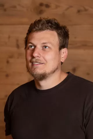 Mitarbeiterportrait / Foto mit braunem T-Shirt oder schwarzem Polo Shirt mit Zimmerei Hirschi Logo. Strahlend und lachend nach oben schauend oder direkt in die Kamera.