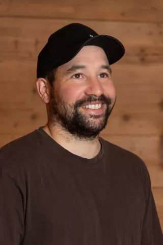 Mitarbeiterportrait / Foto mit braunem T-Shirt oder schwarzem Polo Shirt mit Zimmerei Hirschi Logo. Strahlend und lachend nach oben schauend oder direkt in die Kamera.