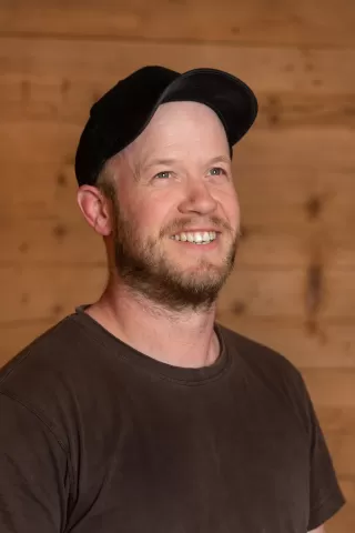 Mitarbeiterportrait / Foto mit braunem T-Shirt oder schwarzem Polo Shirt mit Zimmerei Hirschi Logo. Strahlend und lachend nach oben schauend oder direkt in die Kamera.