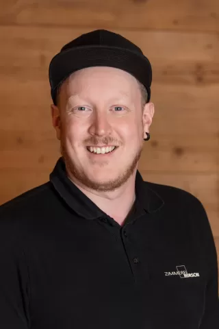 Mitarbeiterportrait / Foto mit braunem T-Shirt oder schwarzem Polo Shirt mit Zimmerei Hirschi Logo. Strahlend und lachend nach oben schauend oder direkt in die Kamera.