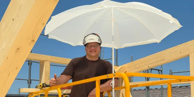 Innenansicht einer im Bau befindlichen Gewerbehalle mit weit gespannter Holzdachkonstruktion aus Brettschichtholz. Sichtbar sind die tragenden Binder, Sparren und die Dachschalung, während eine Hebebühne auf der Holzdecke den Baufortschritt der Umnutzung dokumentiert.