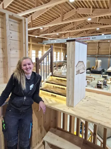 Eine Lernende im Holzbau lehnt lächelnd an einem Holzgeländer und steht neben einem beleuchteten Treppenmodell mit der Aufschrift ‚Treppenmodell, 3. Lehrjahr, 2026‘. Die Szene befindet sich in einer grossen, modernen Zimmerei-Werkhalle mit sichtbarer Holzkonstruktion, Dachbalken und verschiedenen Holzbauelementen im Hintergrund.