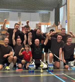 Eine Unihockey-Mannschaft feiert gemeinsam mit einem Kind und einem Torhüter auf dem Hallenboden einen Turniersieg. Vor der Gruppe stehen ein Pokal und zwei Getränkepreise.