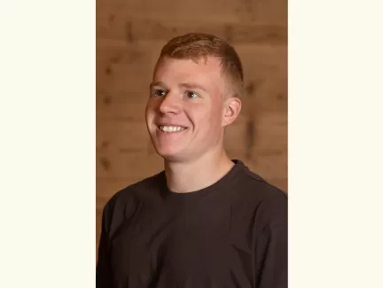 Porträt eines jungen Mannes mit kurzem, hellbraunem Haar, der lächelnd leicht nach rechts blickt. Er trägt ein dunkles T-Shirt, im Hintergrund ist eine warme Holzverkleidung zu sehen.
