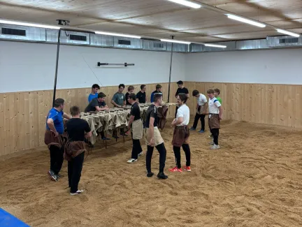 Innenaufnahme einer grossen Halle mit Holzdecke und Holzverkleidung an den Wänden. Mehrere junge Männer stehen in Arbeitskleidung auf einem mit Sägemehl bedeckten Boden und arbeiten gemeinsam an Holzbänken. Die Szene vermittelt eine handwerkliche Ausbildungs- oder Trainingssituation in einer Werk- oder Übungshalle.