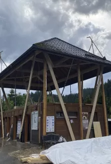 Modernes Holzgebäude mit markantem, erhöhtem Satteldach und offener Konstruktion, aufgenommen an einem bewölkten Tag inmitten einer grünen Hügellandschaft.