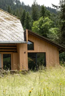 Aussenansicht eines umgebauten Bauernhauses in Holzbauweise mit traditionellem Satteldach, teilweise mit Moos bewachsen. Der Anbau aus hellem Holz fügt sich an den bestehenden Bau an. Im Vordergrund eine Wiese mit hohem Gras, im Hintergrund ein bewaldeter Hang.