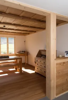 Innenansicht eines wohnlichen Ess- und Arbeitsbereichs in Holzbauweise. Sichtbare Holzbalken an der Decke, ein massiver Holztisch mit Bänken sowie ein Holzschreibtisch stehen in einem hellen Raum mit Holzboden. Tageslicht fällt durch ein Fenster ein und betont die warme, natürliche Atmosphäre sowie die handwerklich ausgeführten Holzdetails.