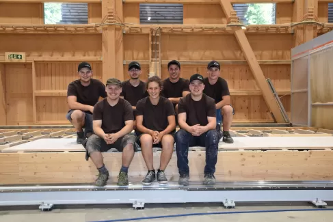 Sieben junge Zimmerleute in Arbeitskleidung posieren lächelnd in einer Werkhalle. Drei Personen sitzen auf einer Holzkonstruktion, vier knien oder stehen dahinter. Alle tragen dunkle T-Shirts und teils Kappen, im Hintergrund ist eine moderne Holzbauhalle sichtbar.