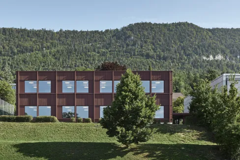 Aussenansicht eines modernen, zweigeschossigen Gebäudes mit dunkelbrauner Fassade und grossen, regelmässig angeordneten Fenstern. Das Gebäude steht in einer grünen Umgebung mit Wiese und Bäumen. Im Hintergrund erhebt sich ein bewaldeter Hügel unter klarem Himmel, was die ruhige, naturnahe Lage des Baus betont.