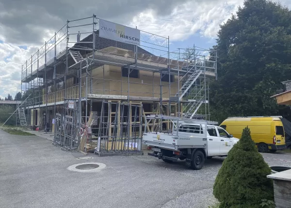 Eingerüsteter Holzrohbau mit Zimmerei-Hirschi-Banner, umgeben von Montagegeräten, Firmenfahrzeug und gelbem Transporter auf dem Baustellengelände.