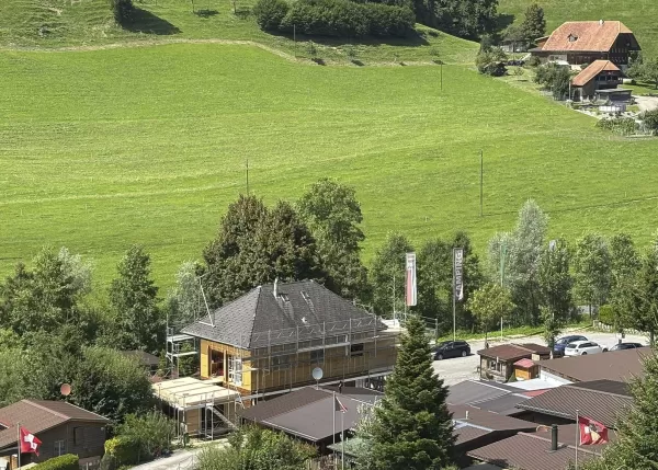 Luftaufnahme eines eingerüsteten Holzbaus inmitten einer kleinen Siedlung, umgeben von grünen Wiesen und Bauernhäusern.