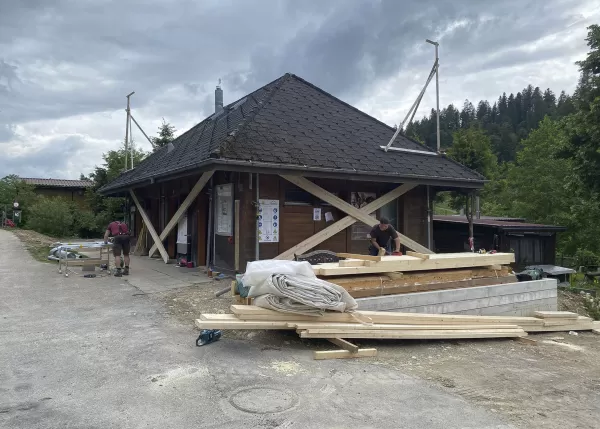 Holzhandwerker bei der Arbeit an einem traditionellen Gebäude mit Schindeldach, umgeben von Baumaterialien, vor bewaldeter Hügellandschaft unter bewölktem Himmel.