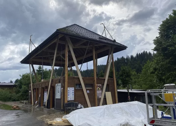 Modernes Holzgebäude mit markantem, erhöhtem Satteldach und offener Konstruktion, aufgenommen an einem bewölkten Tag inmitten einer grünen Hügellandschaft.
