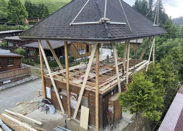Holzgebäude im Umbau mit freigelegtem Deckengerüst, gestützt von hohen Holzbalken unter einem großen Satteldach, umgeben von Chalets und grüner Landschaft.