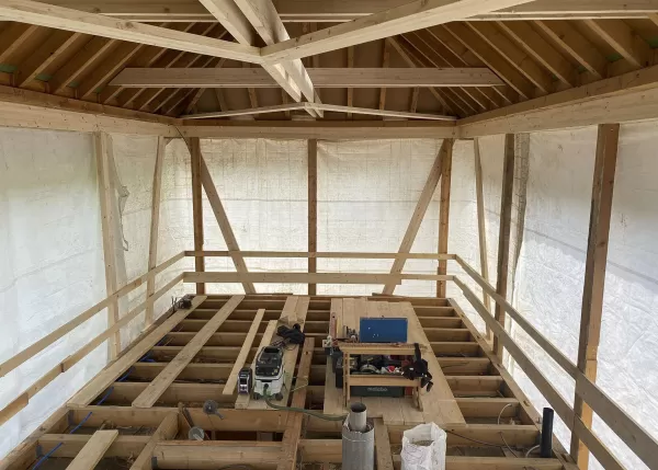 Innenansicht eines Holzbaus im Rohbau mit offenem Boden- und Dachstuhl, Arbeitsgeräten und provisorischer Verkleidung aus Folie.