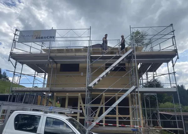 Zwei Personen stehen auf einem eingerüsteten Holzrohbau unter dem Banner von Zimmerei Hirschi, mit einer kleinen Richtbaum-Tanne auf dem Dach.