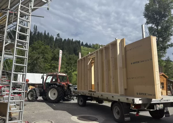 Holzwandelement auf einem Anhänger der Zimmerei Hirschi, gezogen von einem Traktor, bereit zum Einsatz auf einer Baustelle im Grünen.