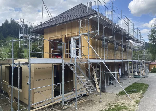 Eingerüsteter Holzbau mit montierten Fassadenelementen und Dach, aufgenommen bei sonnigem Wetter inmitten grüner Landschaft