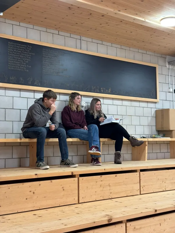 Innenaufnahme einer grossen Halle mit Holzdecke und Holzverkleidung an den Wänden. Mehrere junge Männer stehen in Arbeitskleidung auf einem mit Sägemehl bedeckten Boden und arbeiten gemeinsam an Holzbänken. Die Szene vermittelt eine handwerkliche Ausbildungs- oder Trainingssituation in einer Werk- oder Übungshalle.