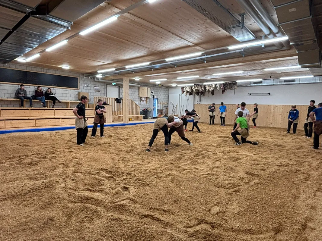 Innenaufnahme einer grossen Halle mit Holzdecke und Holzverkleidung an den Wänden. Mehrere junge Männer stehen in Arbeitskleidung auf einem mit Sägemehl bedeckten Boden und arbeiten gemeinsam an Holzbänken. Die Szene vermittelt eine handwerkliche Ausbildungs- oder Trainingssituation in einer Werk- oder Übungshalle.