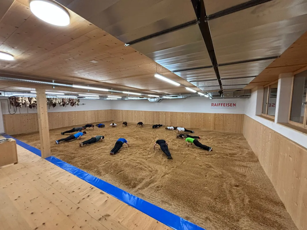Innenaufnahme einer grossen Halle mit Holzdecke und Holzverkleidung an den Wänden. Mehrere junge Männer stehen in Arbeitskleidung auf einem mit Sägemehl bedeckten Boden und arbeiten gemeinsam an Holzbänken. Die Szene vermittelt eine handwerkliche Ausbildungs- oder Trainingssituation in einer Werk- oder Übungshalle.