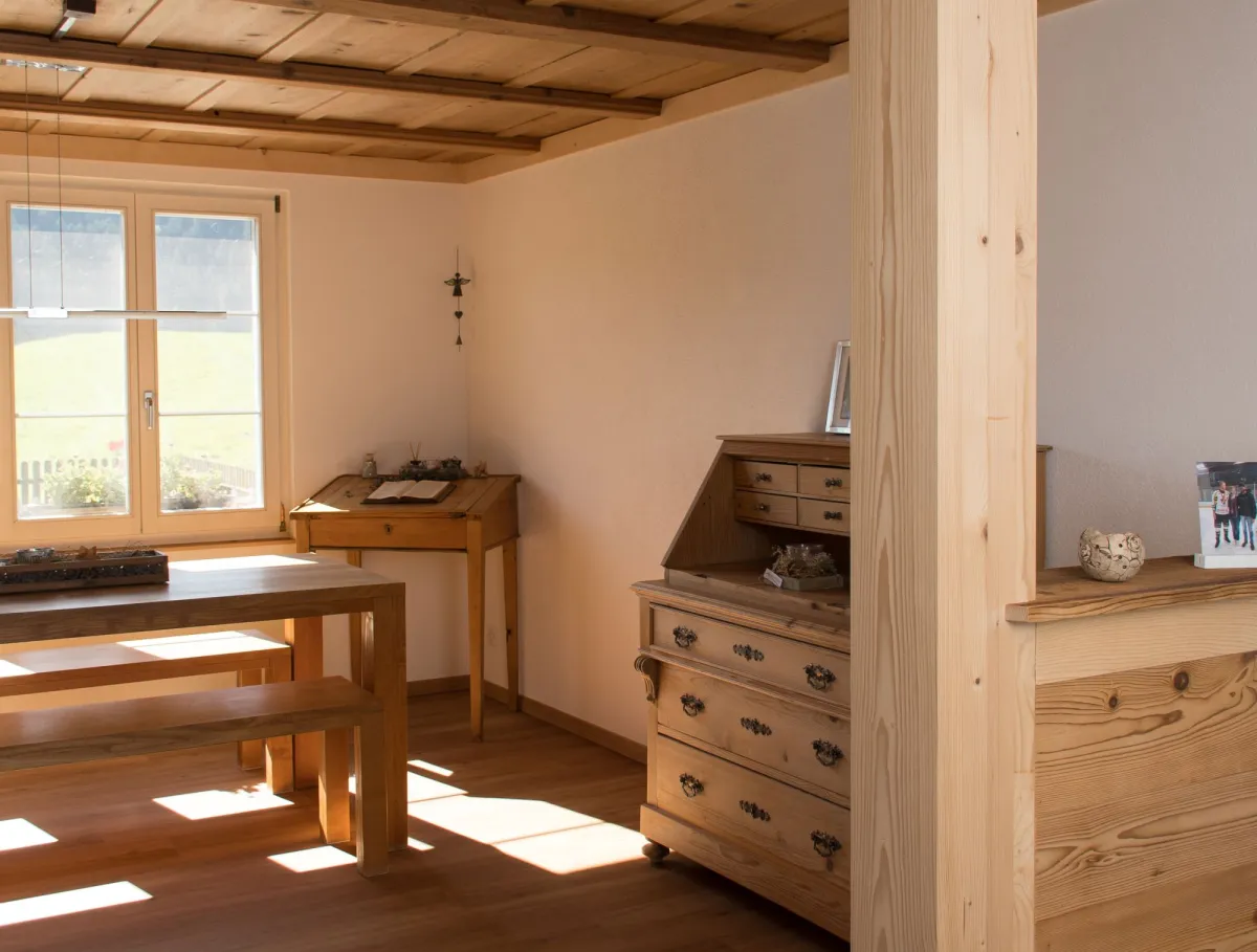 Innenansicht eines wohnlichen Ess- und Arbeitsbereichs in Holzbauweise. Sichtbare Holzbalken an der Decke, ein massiver Holztisch mit Bänken sowie ein Holzschreibtisch stehen in einem hellen Raum mit Holzboden. Tageslicht fällt durch ein Fenster ein und betont die warme, natürliche Atmosphäre sowie die handwerklich ausgeführten Holzdetails.