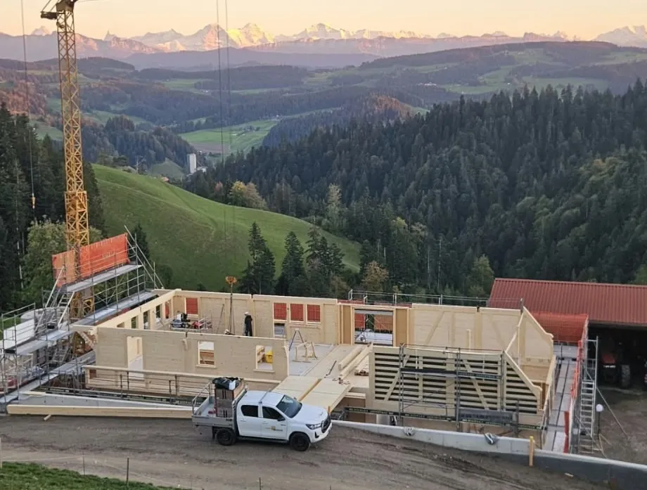 Holzbau im Rohbau mit Baukran an einem grünen Hang, mit bewaldeten Hügeln und schneebedeckten Bergen im Hintergrund.