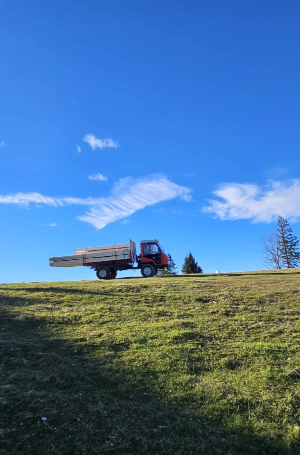 Montagearbeiten an einer Holzplattform vor einem traditionellen Gebäude auf einer Anhöhe. Ein Traktor mit Hebevorrichtung hebt einen langen Holzbalken an, während zwei Mitarbeitende der Zimmerei Hirschi AG den Balken ausrichten.