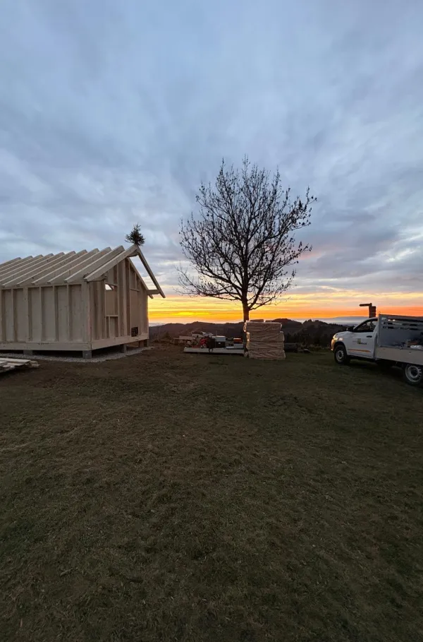 Montagearbeiten an einer Holzplattform vor einem traditionellen Gebäude auf einer Anhöhe. Ein Traktor mit Hebevorrichtung hebt einen langen Holzbalken an, während zwei Mitarbeitende der Zimmerei Hirschi AG den Balken ausrichten.