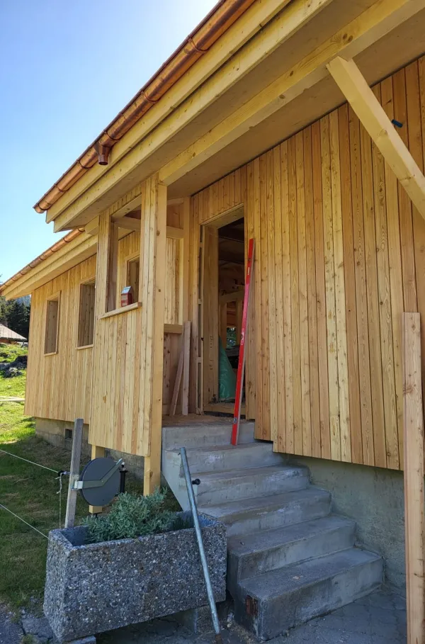 Holzhaus im Bau mit natürlicher Holzfassade inmitten einer Waldlandschaft. Das eingeschossige Gebäude steht auf einem gepflegten Grundstück mit Wiese und Zufahrtsweg. Die Fensteröffnungen sind bereits erstellt, während der Innenausbau noch im Gange ist. Im Hintergrund befinden sich weitere Holzbauten sowie hohe Nadelbäume, die das Gebäude umgeben. Die Aufnahme zeigt die harmonische Einbindung des Holzbaus in die natürliche Umgebung.