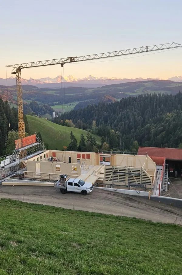 Holzbau im Rohbau mit Baukran an einem grünen Hang, mit bewaldeten Hügeln und schneebedeckten Bergen im Hintergrund.