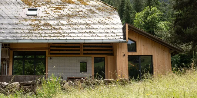 Aussenansicht eines umgebauten Bauernhauses in Holzbauweise mit traditionellem Satteldach, teilweise mit Moos bewachsen. Der Anbau aus hellem Holz fügt sich an den bestehenden Bau an. Im Vordergrund eine Wiese mit hohem Gras, im Hintergrund ein bewaldeter Hang.