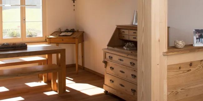 Innenansicht eines wohnlichen Ess- und Arbeitsbereichs in Holzbauweise. Sichtbare Holzbalken an der Decke, ein massiver Holztisch mit Bänken sowie ein Holzschreibtisch stehen in einem hellen Raum mit Holzboden. Tageslicht fällt durch ein Fenster ein und betont die warme, natürliche Atmosphäre sowie die handwerklich ausgeführten Holzdetails.