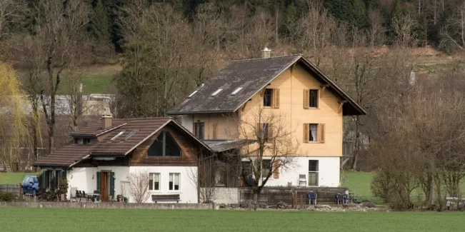 Referenzprojekt der Zimmerei Hirschi AG: Wohnhaus mit umfassend erneuerter Holzfassade, eingebettet in die typische Landschaft des Emmentals mit Wiesen und bewaldetem Hang.