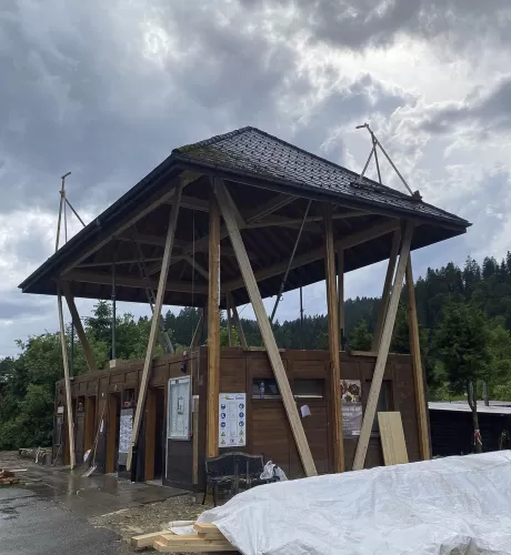 Modernes Holzgebäude mit markantem, erhöhtem Satteldach und offener Konstruktion, aufgenommen an einem bewölkten Tag inmitten einer grünen Hügellandschaft.