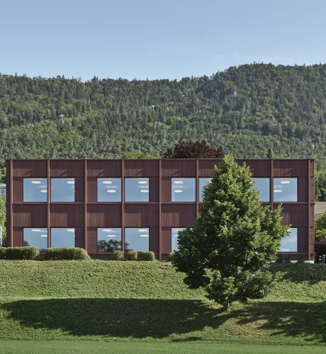 Aussenansicht eines modernen, zweigeschossigen Gebäudes mit dunkelbrauner Fassade und grossen, regelmässig angeordneten Fenstern. Das Gebäude steht in einer grünen Umgebung mit Wiese und Bäumen. Im Hintergrund erhebt sich ein bewaldeter Hügel unter klarem Himmel, was die ruhige, naturnahe Lage des Baus betont.