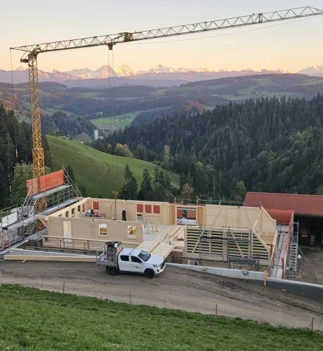 Holzbau im Rohbau mit Baukran an einem grünen Hang, mit bewaldeten Hügeln und schneebedeckten Bergen im Hintergrund.