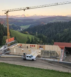 Holzbau im Rohbau mit Baukran an einem grünen Hang, mit bewaldeten Hügeln und schneebedeckten Bergen im Hintergrund.
