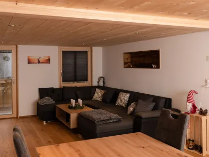 Innenansicht eines gemütlichen Wohnzimmers mit hellem Holzboden und sichtbarer Holzdecke. An der Wand hängt ein Fernseher über einem niedrigen Sideboard. Mehrere Holzfenster geben den Blick in eine bewaldete Landschaft frei. Rechts führt eine Glastür in einen angrenzenden Raum, der Wohnbereich ist warm und wohnlich gestaltet.