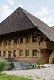 Aussenansicht eines traditionellen Bauernhauses in Holzbauweise mit grossem, steilem Satteldach und holzverschalter Fassade. Das Gebäude verfügt über mehrere Fenster, einen überdachten Bereich sowie eine Aussentreppe. Vor dem Haus befinden sich Sitzgelegenheiten, Pflanzen und ein Kiesplatz, eingebettet in eine ländliche Umgebung mit Hügeln und Bäumen.