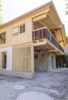 einblicke in ein holzbauprojekt in gunten: die aussenansicht zeigt ein haus mit heller holzfassade und balkon, eingebettet zwischen bäumen und einem steinmäuerchen. im inneren prägen helle holzdecken und sichtbare balken, grosse fenster und warme holzoberflächen die räume. zu sehen sind ein grosszügiger wohnbereich mit zugang zum balkon und ausblick ins grüne, eine holzküche mit arbeitsinsel sowie ein badezimmer mit rundem spiegel und aufgesetztem waschbecken. helles ziegelmauerwerk und sorgfältig erhaltene