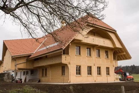 Ein neu erbautes Holzhaus mit roten Ziegeldächern und traditioneller Architektur steht auf einem ländlichen Grundstück; im Vordergrund ist Erde, im Hintergrund sind Wiesen und Bäume zu sehen.